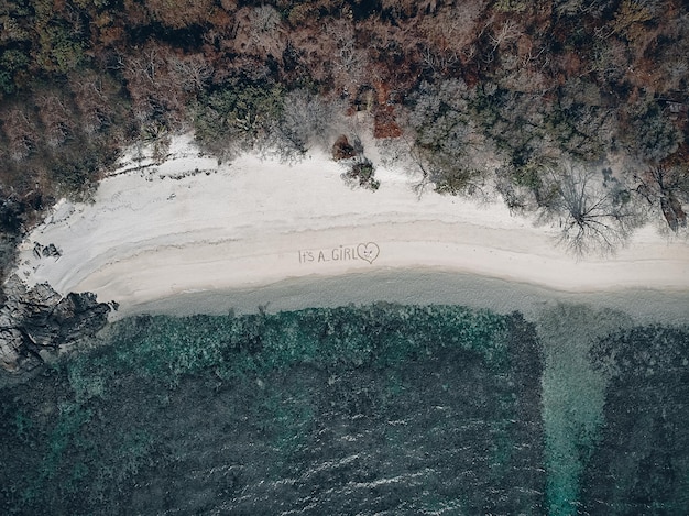 Happy future parents lying on the beach by the turquoise waters. Heart and phrase "It's a girl" written on the sand; drone view.