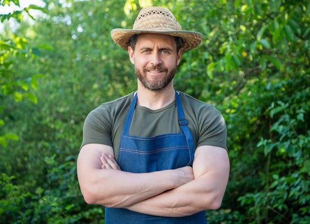 Foto uomo felice del giardiniere che sorride nel grembiule da giardinaggio e nel cappello degli agricoltori che tiene le braccia incrociate in giardino