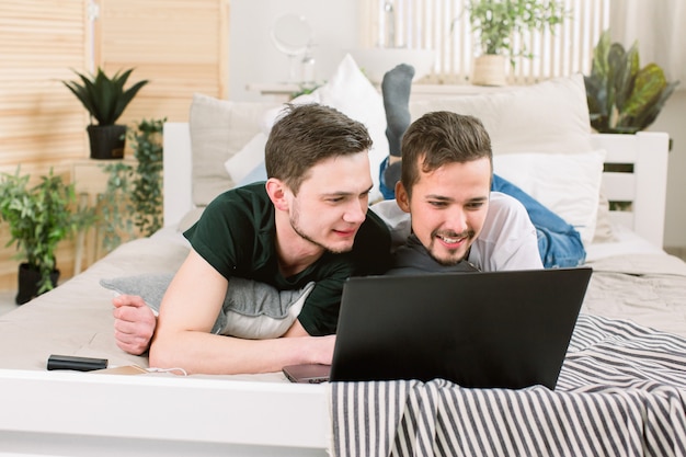 Happy gay couple lying on bed with laptop at home