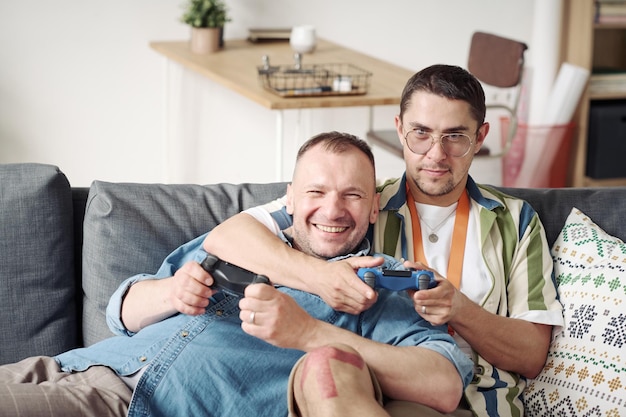 Happy gay couple using joysticks to play game console together while sitting on sofa in living room