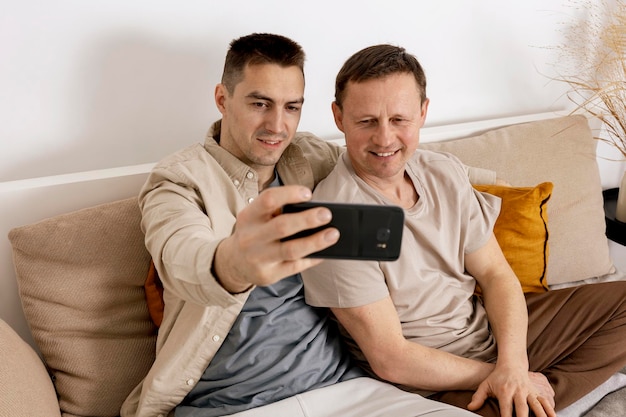 캐주얼한 옷을 입은 행복한 게이 커플은 집에서 함께 시간을 보내고 스마트폰으로 셀카를 찍습니다. 동성애 관계와 대안적인 사랑 아늑한 인테리어