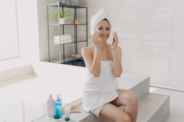 Happy girl applying face lotion with cotton disks Skin purifying and spa procedures at home