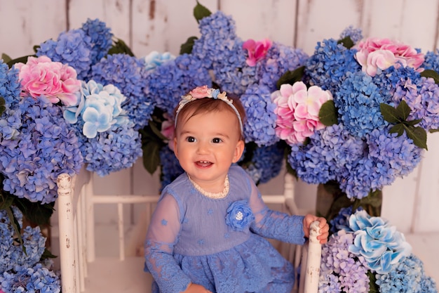 Happy girl in blue dress sits on bed with blue and pink hydrangea flowers