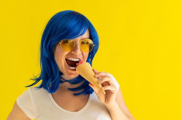 Happy girl in a blue wig and sunglasses eating ice cream cone on a yellow background Emotional excited woman with colored hair is enjoying a cooling gelato on a hot summer day in the studio