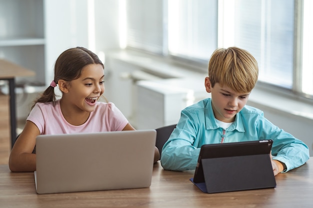 Photo the happy girl and a boy with gadgets sitting at the table