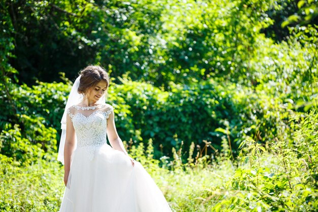 長い白いウェディングドレスと自然の緑の公園で渦巻くベールの幸せな女の子の花嫁。