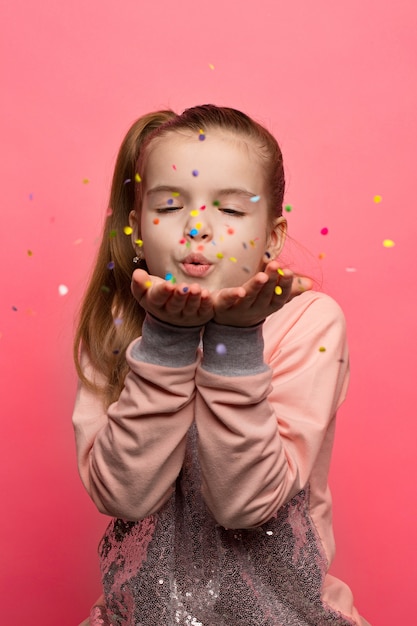 Happy girl celebrating on a pink background. Blows up multicolored confetti