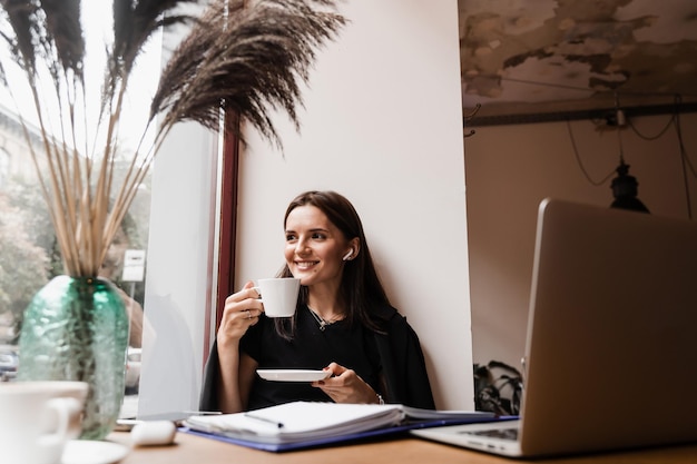 Happy girl chatting with family and friends via video call in loft cafe Successful business woman with laptop works remotely and laughs with colleagues in online video meeting