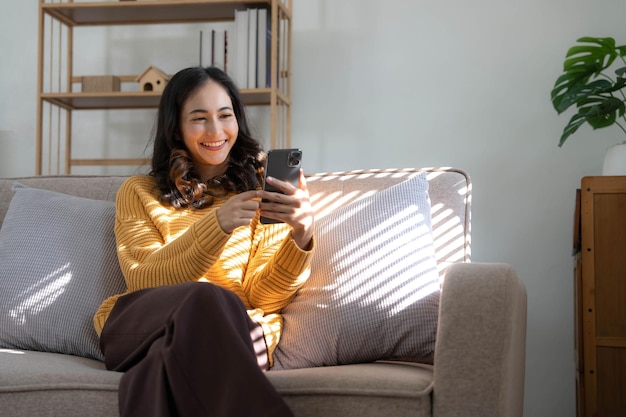 Happy girl checking social media holding smartphone at home Smiling young latin woman using mobile phone app playing game shopping online ordering delivery relax on sofa