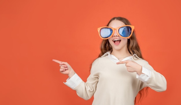 Happy girl child in big funny sunglasses pointing fingers aside brown background copy space