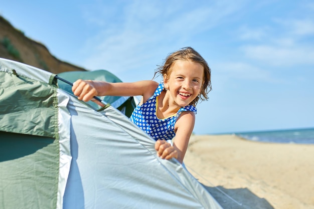 A happy girl child looks out of a tent on the seashore