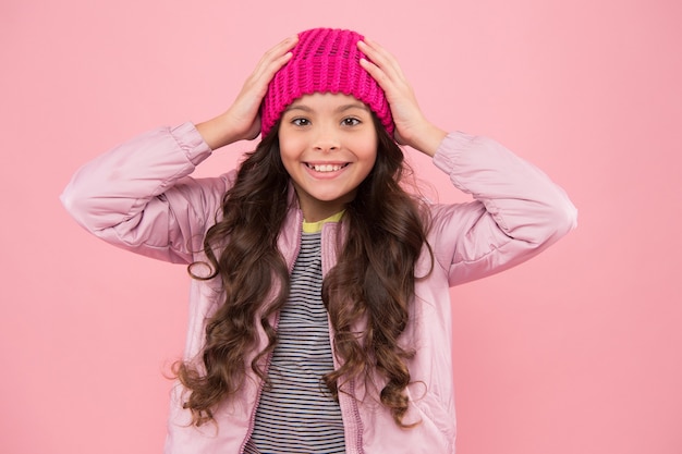 Happy girl child smile with beauty look and long brunette hair fixing fashion beanie hat on head pink background, autumn.