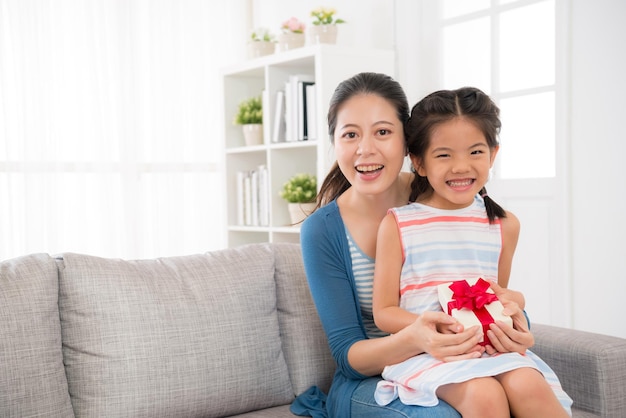 happy girl children and mother hold mother's day gift sitting on the sofa in the living room face the camera together smiling to take pictures commemorate the special holiday with copysapce.