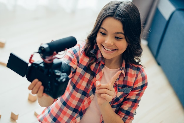 Ragazza felice che conta e punta il dito verso l'alto mentre tiene una moderna fotocamera con microfono e si registra