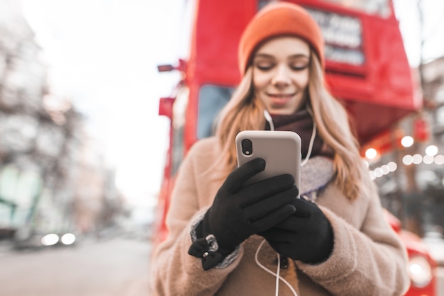 Happy girl dressed in warm clothes standing in the street, listening to music in the headphones and using a smartphone in mittens