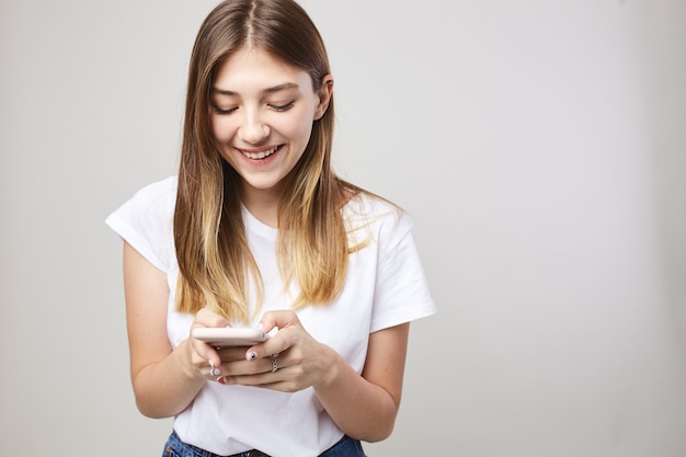 Happy girl dressed in a white tshirt smiles and uses mobile phone on a white background