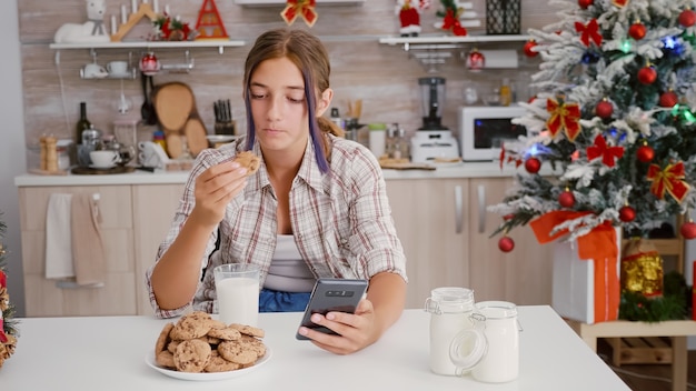 スマートフォンでブラウジングクリスマス装飾キッチンのテーブルに座って冬の休日を楽しんで幸せな女の子