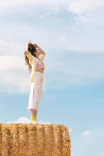 Happy girl enjoys fresh air in the countryside Young beautiful woman with long hair stands on haystack against blue sky