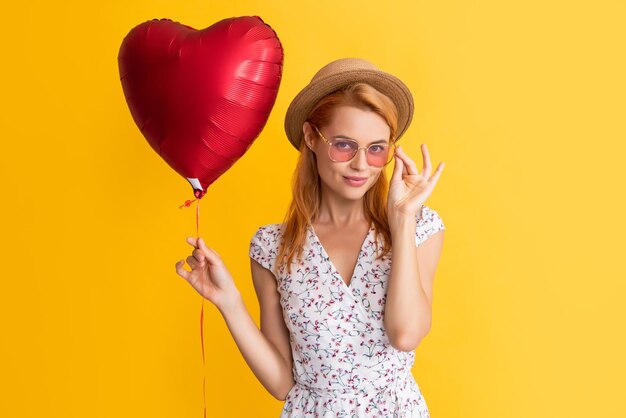 La ragazza felice in occhiali tiene il palloncino del cuore d'amore su sfondo giallo