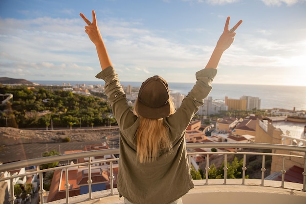 Happy girl from the balcony makes the V sign with her hands and looks at the city and the ocean at sunset