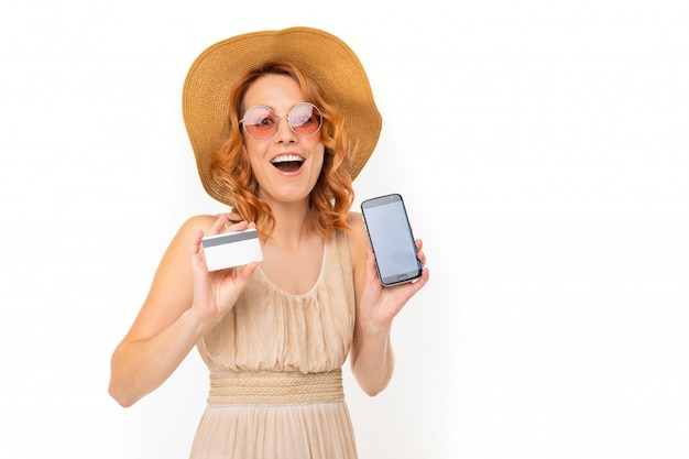 happy girl in a hat dressed in a summer dress with a phone and a credit card with a layout on a white wall