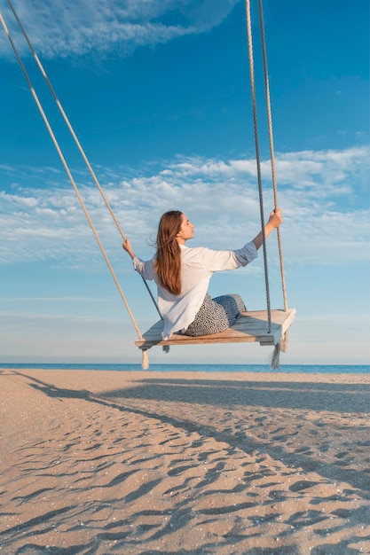 Foto ragazza felice divertirsi oscillando in alto in mezzo all'aria giovane donna in camicia bianca su corda oscillare su cielo blu e fondo mare