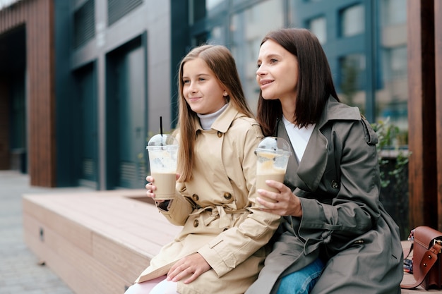 happy girl and her mother in trenchcoats having milk cocktails while relaxing in urban environment
