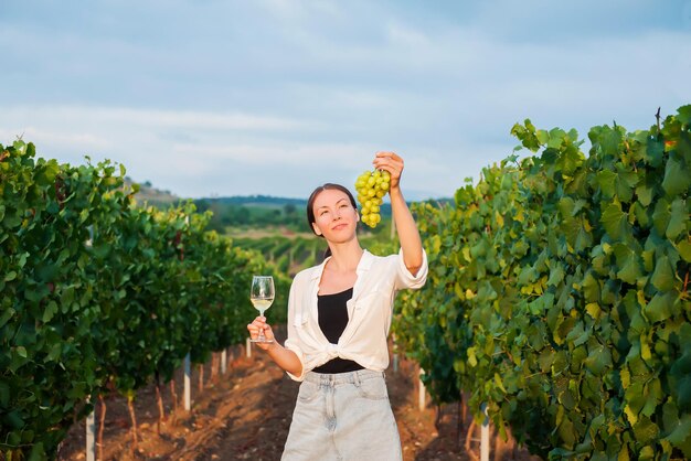 Photo happy girl holding a bunch of grapes and a glass of white wine on the background of a vineyard in the evening at sunset