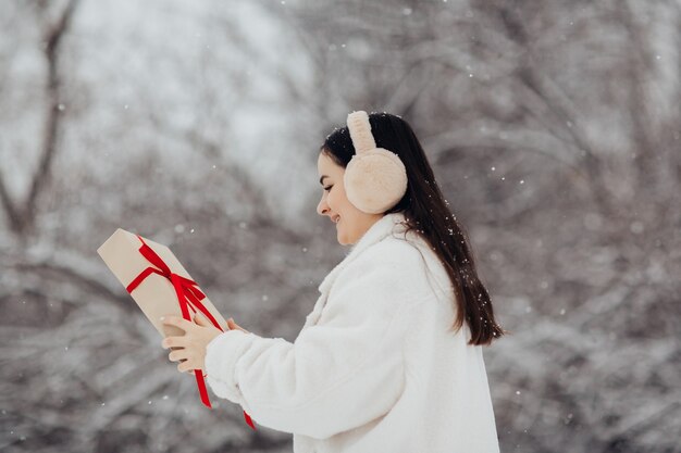 Happy girl holding a gift box with a red ribbon on a snowy winter day for Valentine's Day.