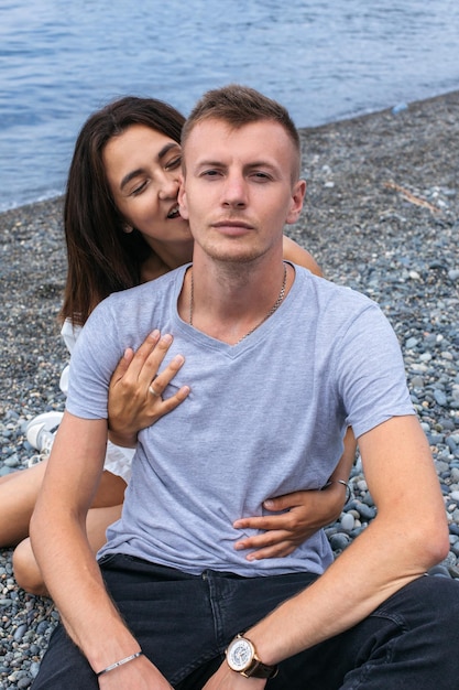 Happy girl hugs her boyfriend from behind and bites his ear on the beach by the sea