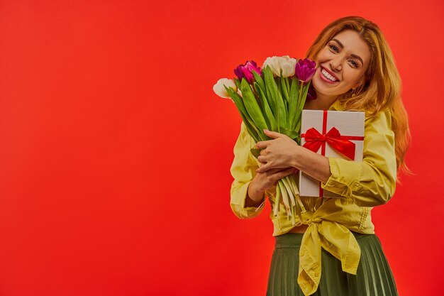 Счастливая девушка в желтой блузке обнимает белую подарочную коробку, перевязанную красной лентой, и букет ярких тюльпанов
