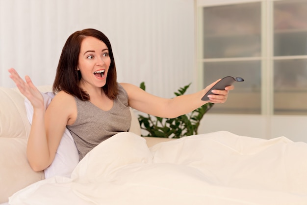 Happy girl is lying on the bed with a remote control for television.