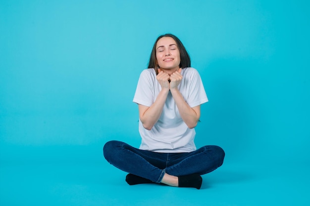Happy girl is wishing by holding hands on chest and sitting on floor on blue background
