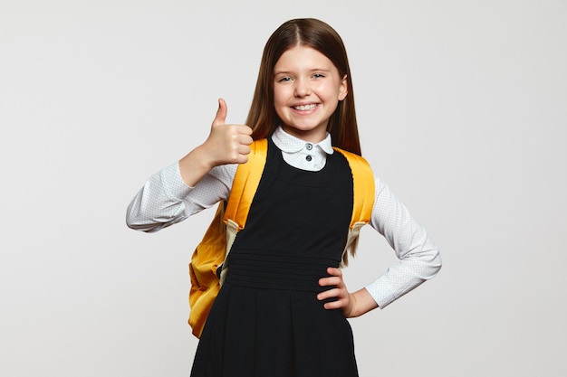 Foto bambino felice della ragazza con lo zaino giallo che indossa l'uniforme scolastica che gesturing il pollice contro il muro bianco