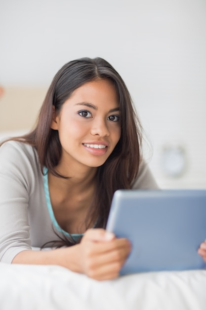 Happy girl lying on bed using her tablet pc smiling at camera