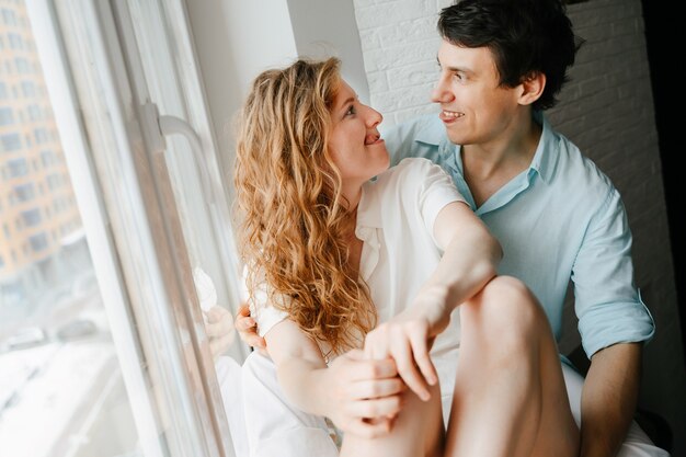 Happy girl and man hugging near window in home. White and blue clothes. Valentine day.