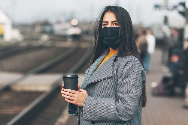 La ragazza felice con una maschera sta aspettando il treno alla stazione con il caffè in mano.