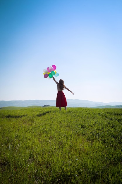 Happy girl in the meadows tuscan with colorful balloons, against the blue sky and green meadow. Tuscany, Italy