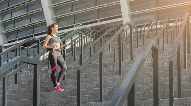 朝の有酸素運動で幸せな女の子。スマートフォンでトレーニング用プレイリストを選択し、水のボトルを保持している若いスポーティな女性