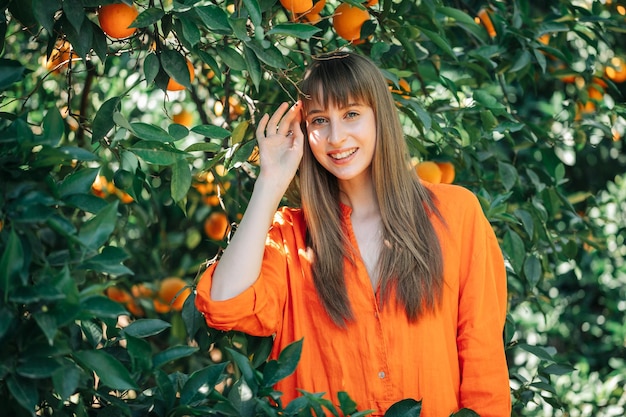 Счастливая девушка в оранжевом платье смотрит в камеру, подняв левую руку в апельсиновом саду