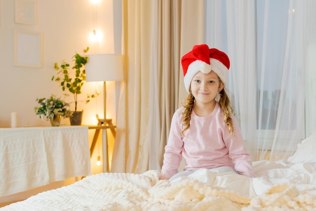 Happy girl in pajamas sits with gift in santa claus hat in room decorated