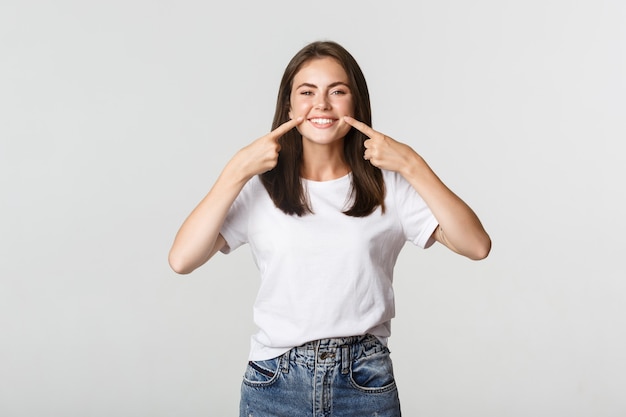 Happy girl pointing fingers at mouth, showing white perfect teeth and beautiful smile.