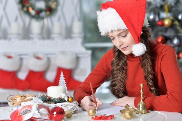 Happy girl in Santa hat preparing for Christmas sitting at the table