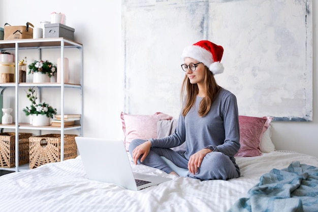 Ragazza felice con cappello da babbo natale che acquista online sul laptop e si siede sul letto accanto