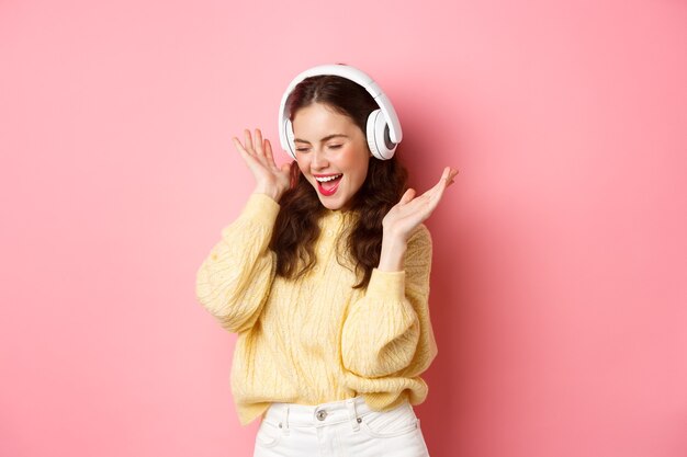 Happy girl singing favorite song, listening to music in wireless headphones, smiling and dancing, standing over pink wall.