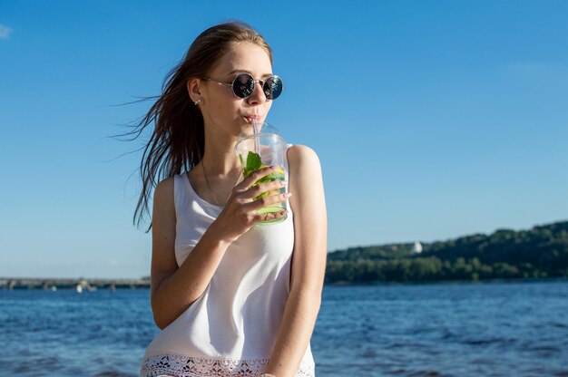 Happy girl sits in blue glasses against the background of the sea and the sky holds a mojito