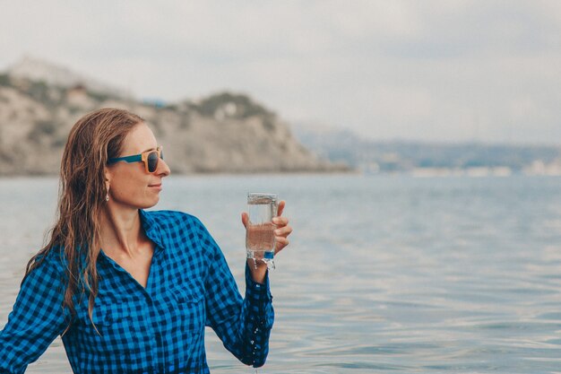 A happy girl sits in the river and drinks water from a glass.