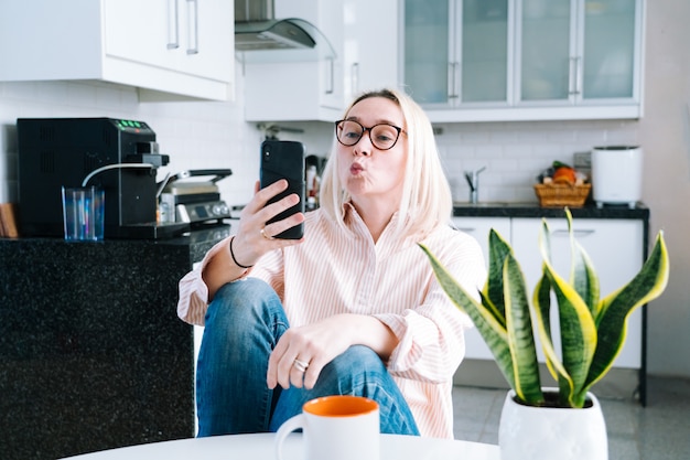 Счастливая девушка сидя дома кухня и держа видеозвонок. Молодая женщина, используя смартфон для видео-звонка с другом или семьей. Запись на вебинар Vlogger. Женщина смотрит в камеру и машет руками приветствия