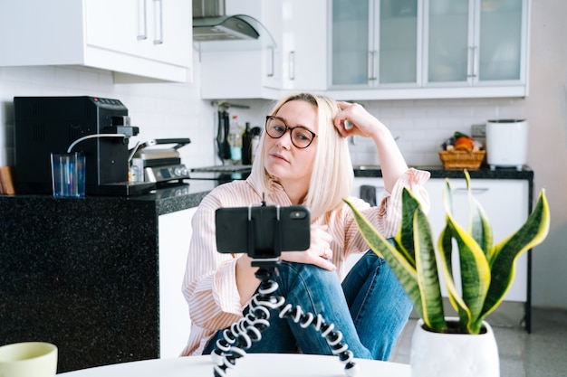 Happy girl sitting at home kitchen and holding videocall. Young woman using smartphone for video call with friend or family. Vlogger recording webinar. Woman looking camera and waving greeting hands