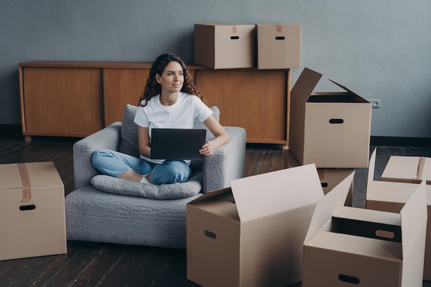 Happy girl sitting at laptop with cardboard boxes dreaming about new first own home relocation